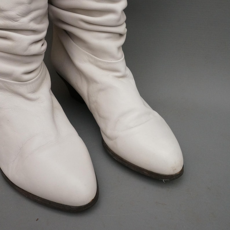 Puede incluir: Un par de botas de cuero blanco roto con un dise&ntilde;o arrugado en la parte superior. Las botas tienen un tac&oacute;n bajo y una punta redondeada. Las suelas son de color marr&oacute;n oscuro. Las botas se muestran sobre un fondo gris.