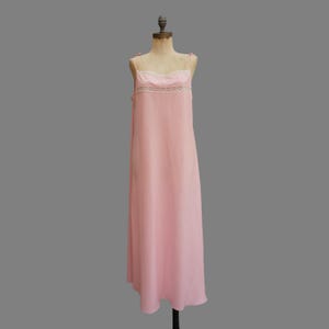 Puede incluir: Un camisón rosa pálido vintage sobre un maniquí. El vestido presenta delicados tirantes finos y un ribete de encaje a lo largo del escote. El diseño largo y fluido sugiere una prenda de dormir o lencería, mostrando un estilo clásico y femenino.