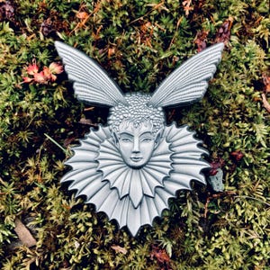 May include: A silver-colored metal brooch featuring a fairy-like face with large, detailed wings and a ruffled collar. The brooch is set against a backdrop of green moss and small plants.