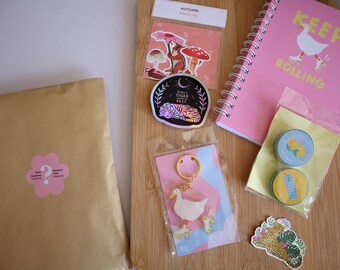 May include: A flat lay of various items on a wooden surface, including a brown paper package with a pink flower sticker, a pink notebook with the text "Keep Rolling", a sticker with a tiger and the text "Even a Tiger needs to rest", a sticker with mushrooms and the text "Autumn Sticker Set", a sticker with a leopard, and a keychain with a white duck on roller skates.