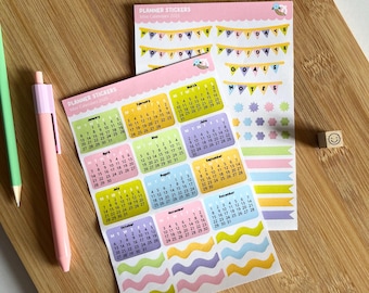 Puede incluir: Un conjunto de pegatinas de planificador coloridas con un mini calendario para 2021, con secciones para vacaciones, objetivos, notas y líneas onduladas decorativas. Las pegatinas están sobre una superficie de madera.
