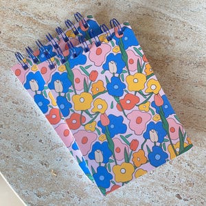 Könnte beinhalten: Drei rosa Spiralblöcke mit einem bunten Blumenmuster mit rosa, blauen, gelben und orangefarbenen Blumen. Die Blöcke haben eine blaue Spiralbindung.