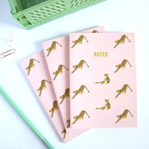 May include: Pale pink notebooks featuring a gold leopard print design and the word 'NOTES' in gold. A green pencil, a green plastic crate, a paperclip, and a drawing pin are visible in the background.