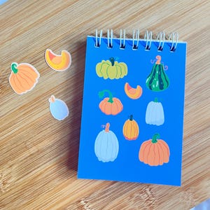 May include: A blue spiral-bound notepad featuring various pumpkin illustrations in orange, green, and light blue. Several pumpkin stickers are scattered around the notepad on a wooden surface. The pumpkins vary in shape and style, perfect for fall-themed crafts.
