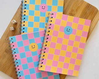 May include: Three spiral bound notebooks with pink and blue checkered covers. Each notebook has a different colored smiley face on the front.