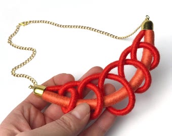 Op de afbeelding: Een rode en oranje statement ketting met een gouden ketting en een geknoopt ontwerp.