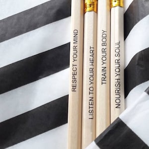 Puede incluir: Cuatro lápices con gomas blancas y bandas metálicas doradas. Cada lápiz tiene una frase inspiradora: "Respeta tu mente", "Escucha a tu corazón", "Entrena tu cuerpo" y "Nutre tu alma". Los lápices están sobre un fondo a rayas blancas y negras.
