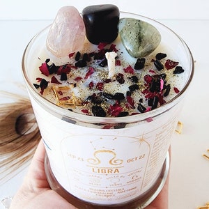 May include: A white candle with a gold label that says "LIBRA" and "SEP 23 - OCT 22". The candle is decorated with black and red glitter, black and pink crystals, and a clear quartz crystal. The candle is in a clear glass jar.