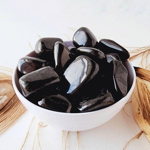 May include: A white bowl filled with black tumbled stones. The stones are smooth and polished, and they have a natural, earthy look.