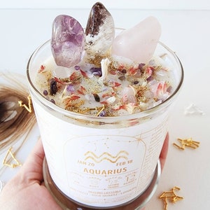 May include: A white candle with a clear glass jar and a gold label that says "Aquarius" with the dates "Jan 20" and "Feb 18". The candle is decorated with crystals, including amethyst, rose quartz, and clear quartz. The candle is surrounded by gold and silver decorative accents.