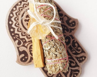 May include: A wooden hand-shaped holder with a mandala design, featuring a bundle of sage smudge sticks, a palo santo stick, and a clear citrine crystal.