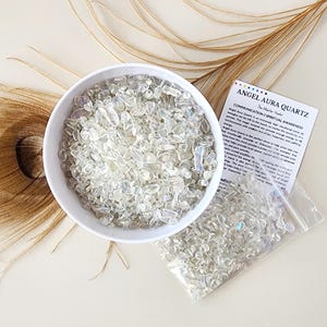 Peut inclure: Un bol en céramique blanc rempli de cristaux de quartz Angel Aura clairs et irisés. Un petit sac en plastique contenant les mêmes cristaux est posé à côté du bol. Une carte avec le texte "Angel Aura Quartz" est également présente. Les cristaux sont sur une surface crème avec une plume.
