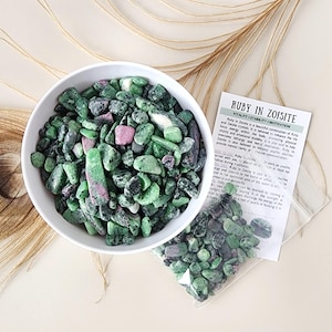 May include: A white bowl filled with green and pink Ruby in Zoisite stones. A small clear bag of stones and a printed card with text are next to the bowl. The background includes a peacock feather and a dried plant stem.
