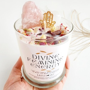 May include: A white candle with a pink rose quartz crystal, gold hamsa hand charm, and dried flowers. The candle label reads "Divine Feminine Energy" and "Light this candle while focusing on what you want to manifest." The label also includes the text "Krisla Art" and "Infused with positive energy & essential oils."