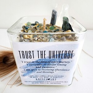May include: A square glass candle filled with white wax, topped with various small stones, crystals, and dried botanicals. The label reads: TRUST THE UNIVERSE, I trust in the flow of life's journey, I surrender to divine timing and guidance.