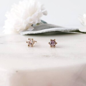 May include: Two gold stud earrings with a prong setting. The earring on the left has a clear stone, while the earring on the right has a light purple stone.