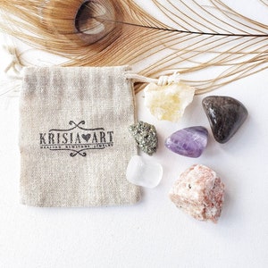 May include: A small burlap pouch with the text "KRISJA HEART HEALING CRYSTALS JEWELLERY" printed on it. The pouch is surrounded by six small crystals: a clear crystal, a purple amethyst, a yellow citrine, a brown crystal, a green crystal, and a pink crystal.