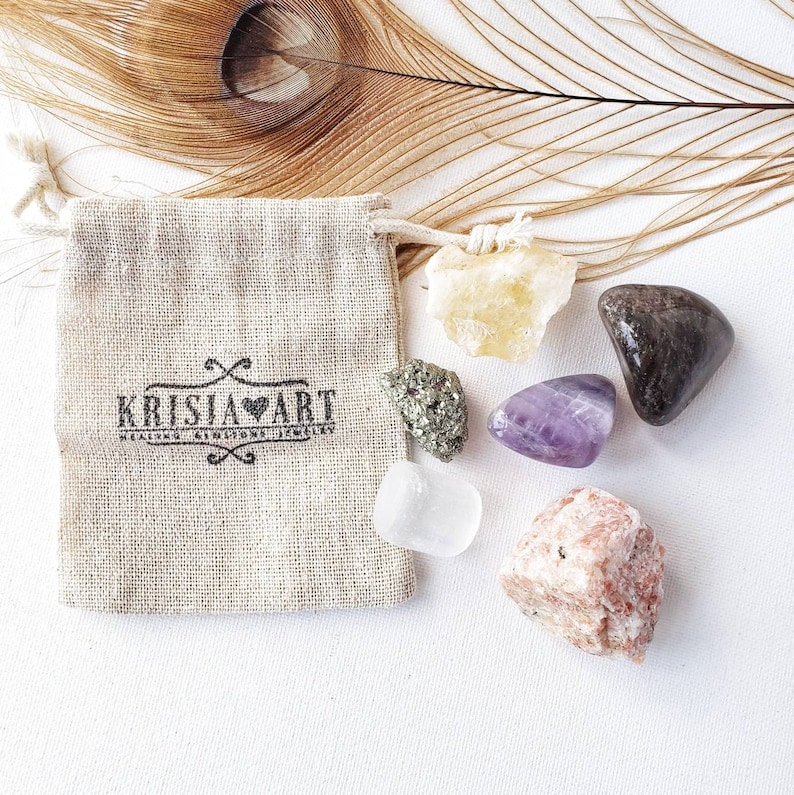 May include: A small burlap pouch with the text "KRISJA HEART HEALING CRYSTALS JEWELLERY" printed on it. The pouch is surrounded by six small crystals: a clear crystal, a purple amethyst, a yellow citrine, a brown crystal, a green crystal, and a pink crystal.
