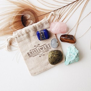 May include: A small pouch with a drawstring closure containing six tumbled crystals: blue lapis lazuli, pink rose quartz, brown tiger's eye, green aventurine, clear quartz, and a dark green stone. The pouch is made of natural fabric and has the text "Krista Art" printed on it.