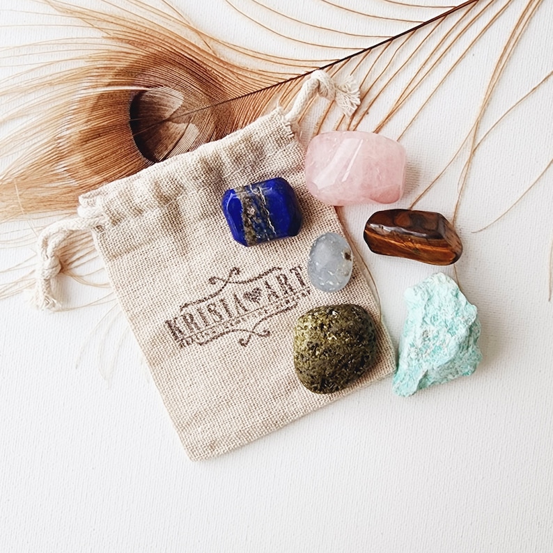 May include: A small pouch with a drawstring closure containing six tumbled crystals: blue lapis lazuli, pink rose quartz, brown tiger's eye, green aventurine, clear quartz, and a dark green stone. The pouch is made of natural fabric and has the text "Krista Art" printed on it.