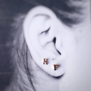 May include: Close-up of a person's ear with two gold initial earrings, one with the letter "K" and the other with the letter "P".
