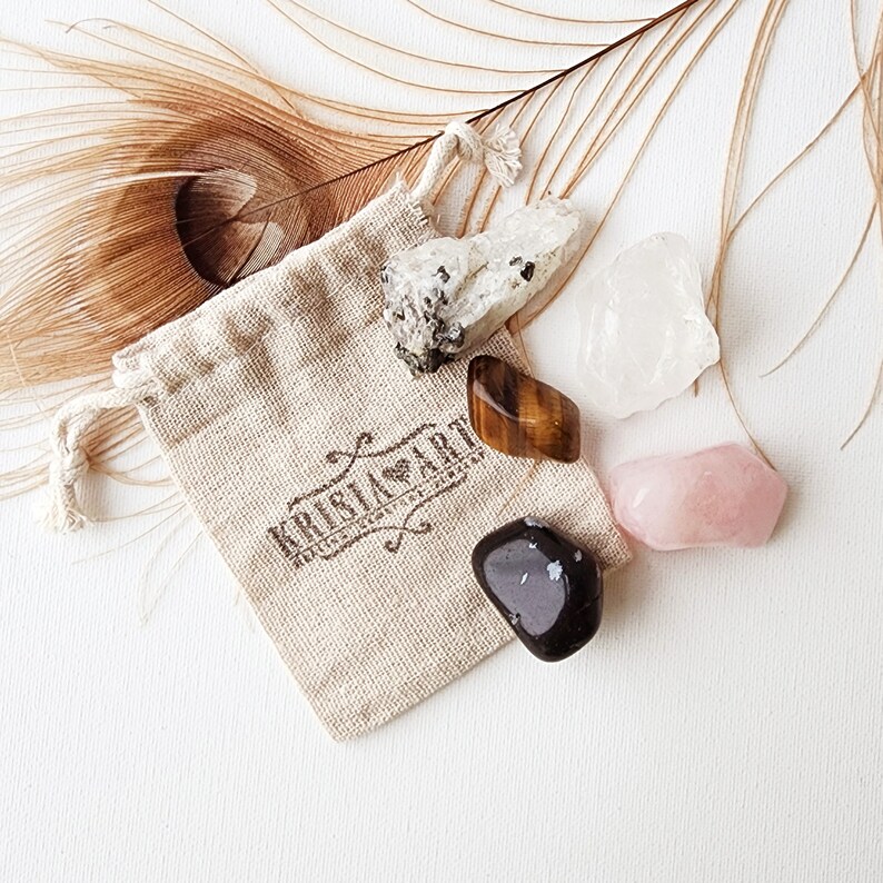May include: A small, beige cloth bag with a drawstring closure, containing five tumbled crystals: clear quartz, tiger's eye, rose quartz, black obsidian, and a white crystal with black flecks. The bag has a brown logo that reads "KRISTAL ART" with a heart symbol.