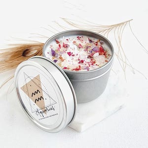 May include: A silver tin candle with a white wax base and pink and purple flower petals and crystals. The label on the tin says "Aquarius" with a stylized astrological symbol.