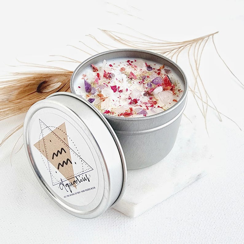 May include: A silver tin candle with a white wax base and pink and purple flower petals and crystals. The label on the tin says "Aquarius" with a stylized astrological symbol.