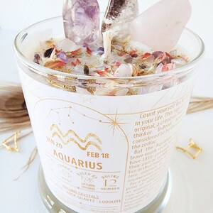 May include: A white candle with a gold label that says "Aquarius" and "Jan 20 - Feb 18". The candle is decorated with pink, purple, and white crystals. The label also includes the ruling crystals, rose quartz and lodalite.