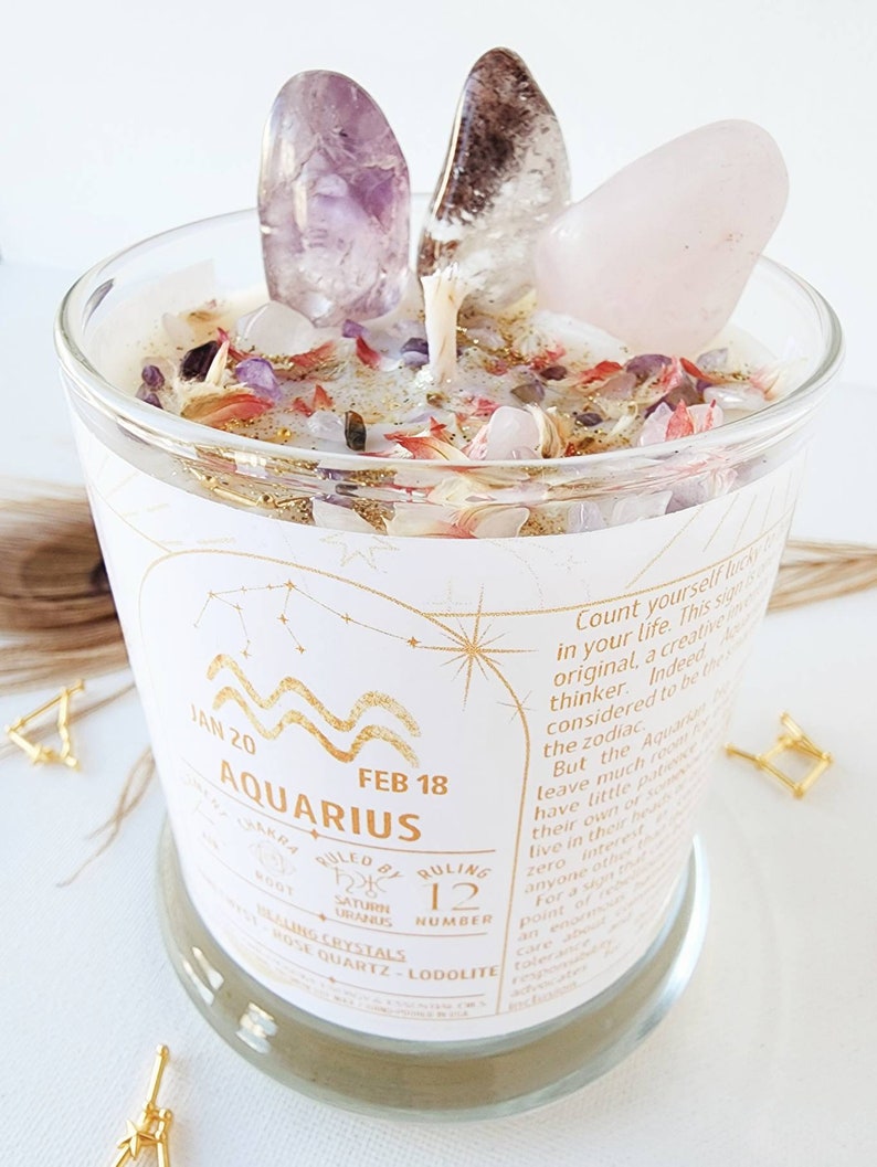 May include: A white candle with a gold label that says "Aquarius" and "Jan 20 - Feb 18". The candle is decorated with pink, purple, and white crystals. The label also includes the ruling crystals, rose quartz and lodalite.