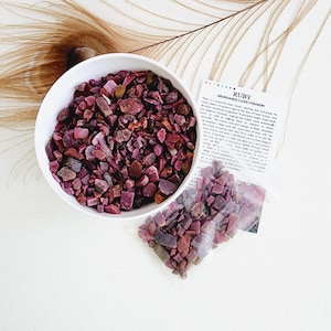 May include: A white bowl and a small plastic bag filled with small, red ruby gemstone chips. The bag has a label that reads "RUBY ABUNDANCE I LOVE PASSION".
