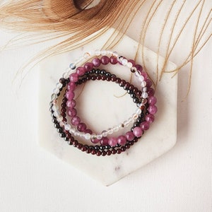May include: Four stacked bracelets made of small, round beads. The bracelets are in shades of purple, white, and black.