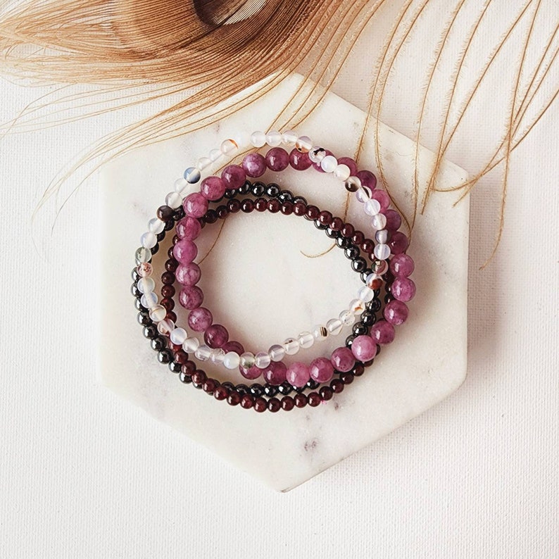 May include: Four stacked bracelets made of small, round beads. The bracelets are in shades of purple, white, and black.