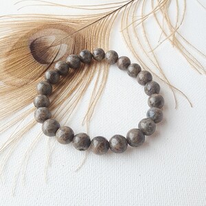 May include: A brown beaded bracelet made of round, speckled stones. The bracelet is lying on a white surface with a brown feather behind it.