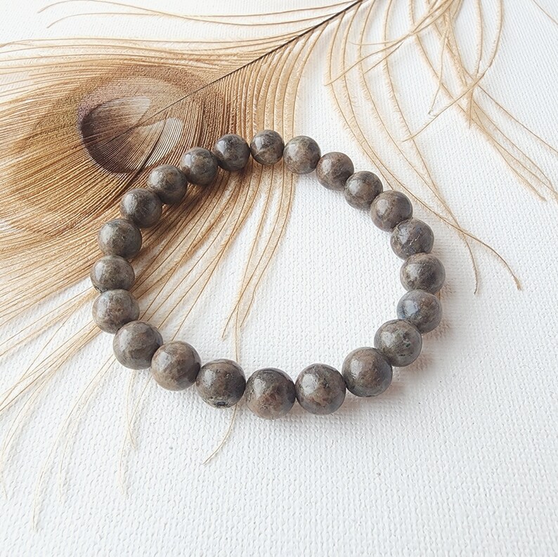 May include: A brown beaded bracelet made of round, speckled stones. The bracelet is lying on a white surface with a brown feather behind it.