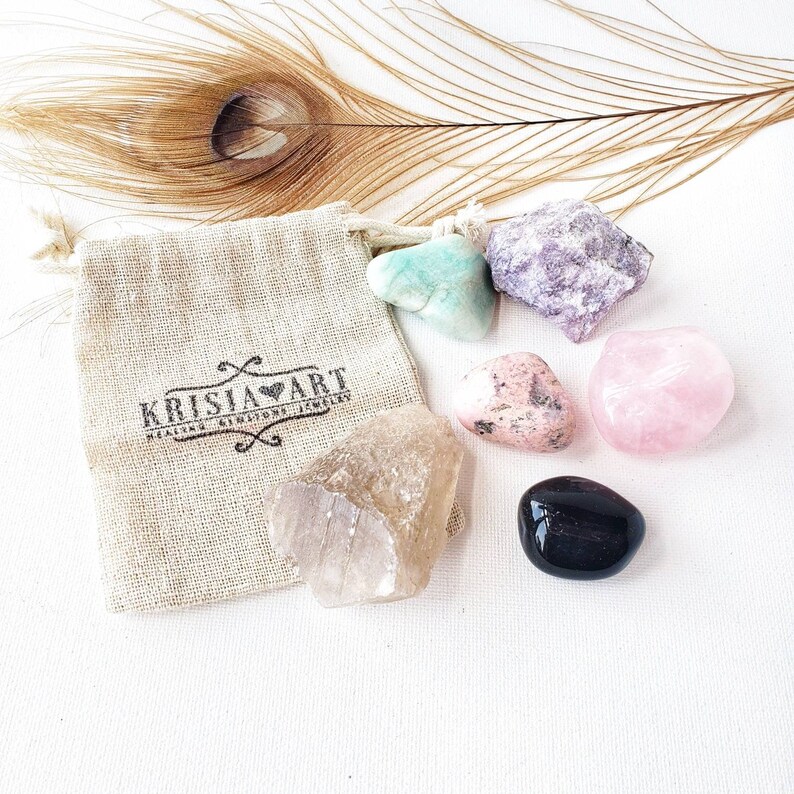 Puede incluir: Un conjunto de siete cristales pulidos en una peque&ntilde;a bolsa de arpillera. Los cristales son de color rosa, verde, morado, transparente y negro. La bolsa tiene el texto "Krisia Art Healing Crystals Jewelry" impreso en ella.