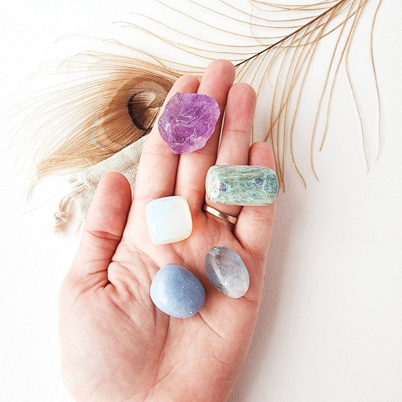 May include: A collection of five tumbled crystals: amethyst, clear quartz, blue lace agate, green kyanite, and a light blue stone. The crystals are arranged in a hand, with the amethyst at the top and the light blue stone at the bottom.