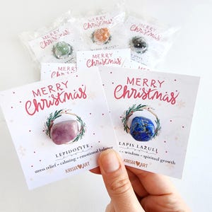 May include: Two white gift tags with the text "Merry Christmas" in red. Each tag features a different crystal, a pink Lepidolite and a blue Lapis Lazuli, with a wreath around them. The text below each crystal describes the crystal's properties.
