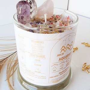May include: A white candle in a glass jar with a gold label. The label has the astrological sign Aquarius, the dates January 20 to February 18, and the words "Element: Air, Chakra: Root, Healing Crystals: Amethyst - Rose Quartz". The candle is surrounded by pink and purple crystals.