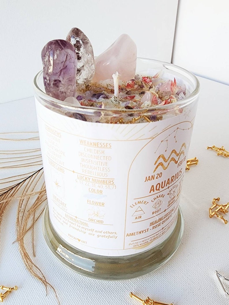 May include: A white candle in a glass jar with a gold label. The label has the astrological sign Aquarius, the dates January 20 to February 18, and the words "Element: Air, Chakra: Root, Healing Crystals: Amethyst - Rose Quartz". The candle is surrounded by pink and purple crystals.