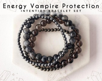 May include: An "Energy Vampire Protection Intention Bracelet Set" is displayed. The set includes three beaded bracelets in shades of gray, brown, and silver. The bracelets are resting on a white, hexagonal tray. The text "Energy Vampire Protection Intention Bracelet Set" is at the top.