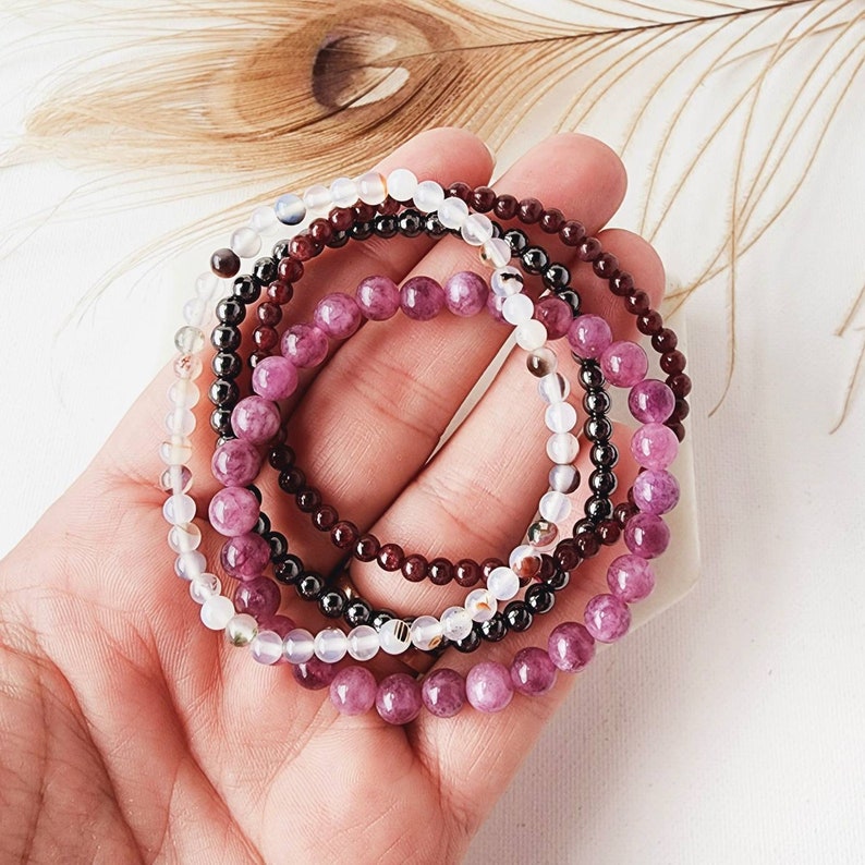 May include: A set of five beaded bracelets in shades of purple, white, and burgundy. The bracelets are stacked on top of each other and are made of small, round beads.