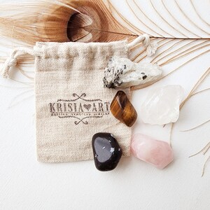 May include: A small pouch with a brown logo that says "Krista Art" contains five tumbled stones: clear quartz, rose quartz, tiger's eye, black obsidian, and a white stone with black flecks.