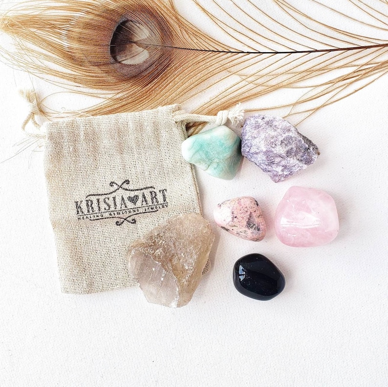 Puede incluir: Un conjunto de seis piedras pulidas en una peque&ntilde;a bolsa de arpillera. Las piedras son de varios colores, incluyendo rosa, verde, negro y morado. La bolsa tiene el texto "Krisia Art Healing Crystal Jewelry" impreso en ella.