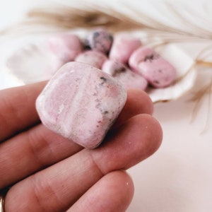 May include: A pink and white tumbled stone with a smooth, polished surface. The stone has a natural, organic shape and is a popular choice for crystal healing.