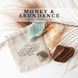 May include: A set of five crystals in a small beige pouch. The crystals are green aventurine, clear quartz, pyrite, tiger's eye, and rose quartz. The pouch has the text "Krisla Art" printed on it. The crystals are labeled as a "Money & Abundance Healing Crystals Set".
