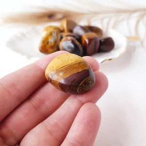 May include: A smooth, polished, brown, yellow and red banded tumbled stone. The stone is being held in a hand.