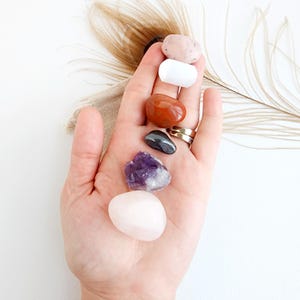 May include: A collection of seven tumbled stones, including rose quartz, amethyst, red jasper, hematite, white selenite, and two pink stones. The stones are arranged in a vertical line in a hand.
