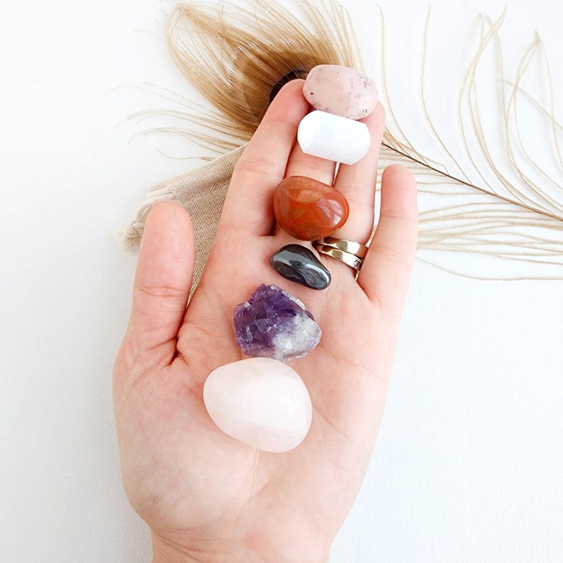 May include: A collection of seven tumbled stones, including rose quartz, amethyst, red jasper, hematite, white selenite, and two pink stones. The stones are arranged in a vertical line in a hand.
