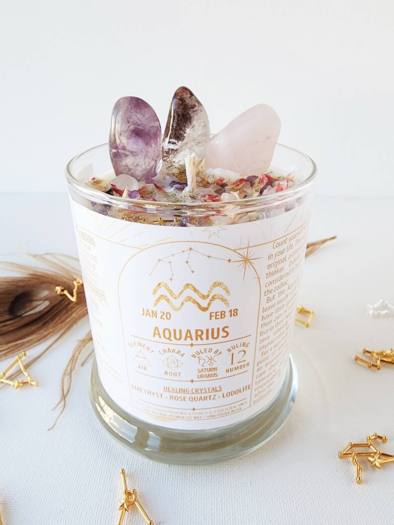 May include: A glass candle jar with a white label featuring the zodiac sign Aquarius, dates January 20 to February 18, and the text "Element: Air, Chakra: Root, Ruled by: Saturn Uranus, Number: 12, Healing Crystals: Amethyst, Rose Quartz, Lodolilte". The jar is filled with a white candle and topped with amethyst, clear quartz, and rose quartz crystals.